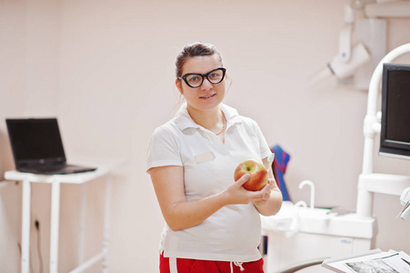 戴眼镜的女牙医的肖像，站在她的牙科办公室里，拿着苹果和牙刷。