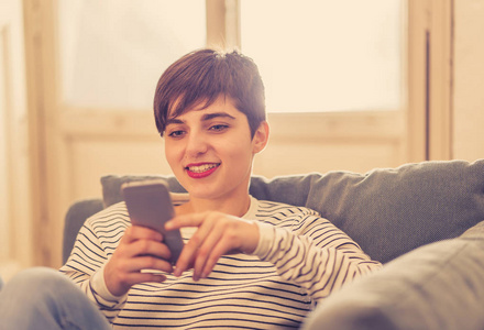 快乐漂亮的年轻白种人女士在智能手机上交谈和检查互联网和社交媒体上的信息，坐在沙发上，在生活方式沟通和人的概念上。