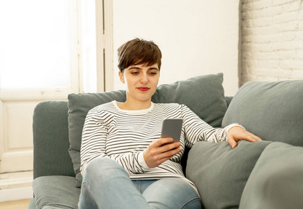 快乐迷人的短发年轻女子在移动智能手机聊天和使用社交媒体应用程序坐在沙发上在网上约会，使用新技术和社交网络概念。