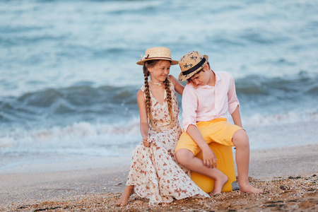 孩子们在海滩上玩和玩。 可爱的青少年。 海滨的浪漫故事。 女孩和男孩坐在海滩上的一个黄色手提箱上。 复古风格