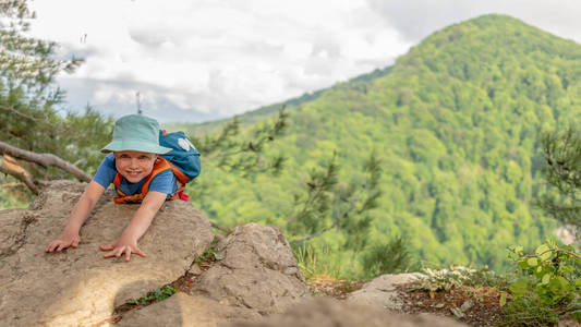 一个带背包的男孩爬上了被绿色森林包围的悬崖顶部。