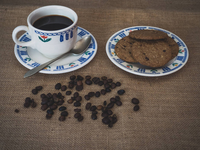 一个瓷质咖啡杯的早餐，一个古色古香的勺子，巧克力饼干和咖啡豆，在一个老式的麻布背景上。