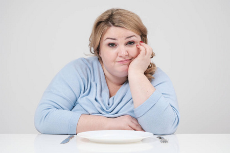 年轻的曲线胖女人穿着休闲的蓝色衣服, 白色背景在桌子前坐在一个空盘子前, 挨饿。饮食和适当的营养