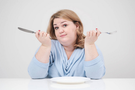 年轻的曲线胖女人穿着休闲的蓝色衣服, 白色背景在桌子前坐在一个空盘子前, 挨饿。饮食和适当的营养