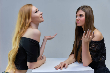 概念肖像，两个可爱的美丽女孩谈论灰色背景在工作室站在桌子前的相机。 他们以不同的姿势表现出不同的情绪。