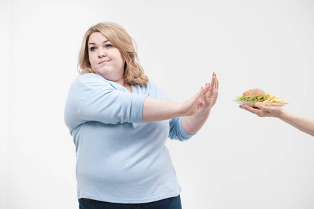 一个年轻的华丽的胖女人穿着休闲的蓝色衣服在白色的背景下拒绝从快餐提供给她, 一个汉堡包和炸薯条。饮食和适当的营养