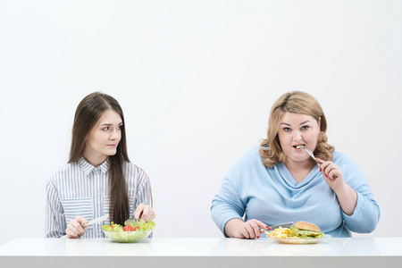苗条的女孩吃健康的食物, 胖女人吃有害的快餐。在白色背景下, 饮食和适当营养的主题