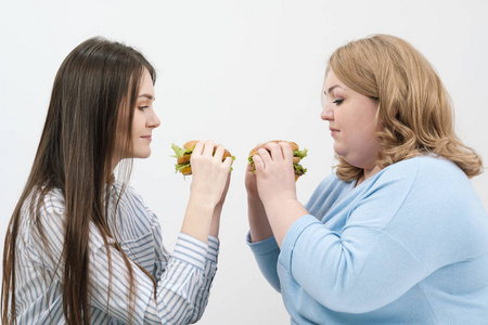两个女孩, 身材苗条, 胖胖的, 金发碧眼的, 黑发的, 吃汉堡包。在白色背景下, 饮食和适当营养的主题