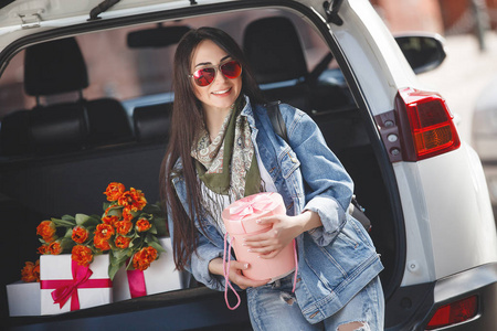 年轻迷人的女人在车里带着礼物盒礼物和鲜花。 春天美丽的女士，带着一束郁金香。 女在汽车里。