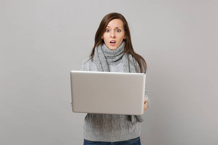 困惑的年轻女子穿着灰色毛衣围巾，工作在笔记本电脑上，电脑孤立在灰色的墙壁背景上。 健康生活方式在线治疗咨询冷季概念。 模拟复制空