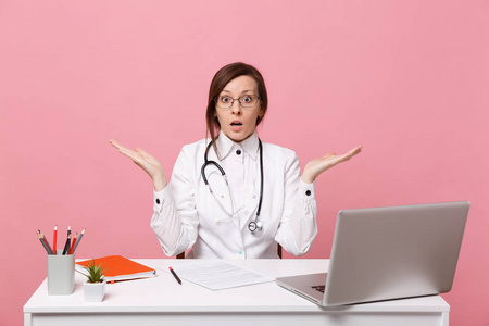 疲惫悲伤的女医生坐在电脑上的办公桌上工作，医院里的医疗文件孤立在粉墙背景上。 穿着医用长袍眼镜的女人听诊器。 保健医学概念
