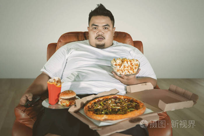 一个肥胖男人在沙发上看电视在家吃垃圾食品的照片