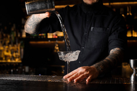 纹身的酒保把一杯酒精饮料从钢制摇床倒入酒吧柜台上的空鸡尾酒玻璃上