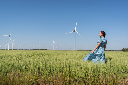 自由的概念。美丽的妇女在蓝色礼服在绿色领域与风发电机涡轮和蓝天背景
