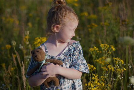 生活方式照片肖像一个女孩在户外的花田向远处看。 在一只针织熊的手中，红色的头发聚集在一个发髻上。