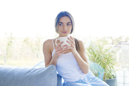 美丽的年轻的年轻女人的肖像照片，她手里拿着杯子，坐在家里的沙发上放松。