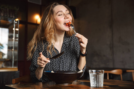 一张兴奋的照片，美丽的年轻漂亮的女人坐在咖啡馆里，在室内吃早餐，吃沙拉