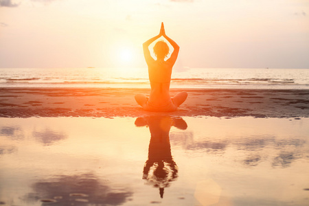 在日落时坐在海滩上的莲花姿势的瑜伽女人