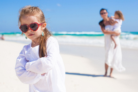 可爱的小女孩和她的妈妈和在地处热带的海滩背景中的小妹妹的画像