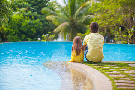 一个年轻的父亲和他可爱的女儿坐在游泳池边的后视图