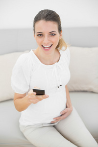 开朗怀孕的妇女抱着她坐在沙发上的智能手机