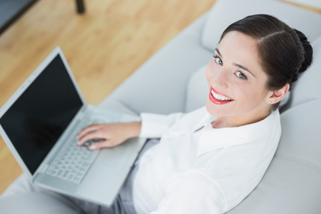 微笑衣冠楚楚的年轻女子在沙发上使用便携式计算机