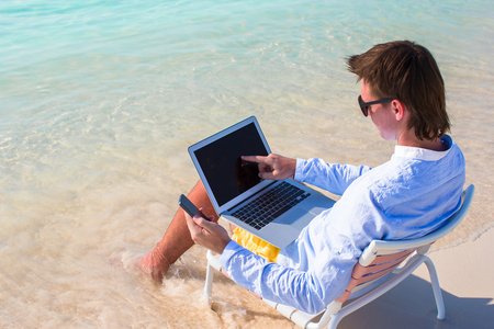 年轻的商人在地处热带的海滩上使用便携式计算机和电话