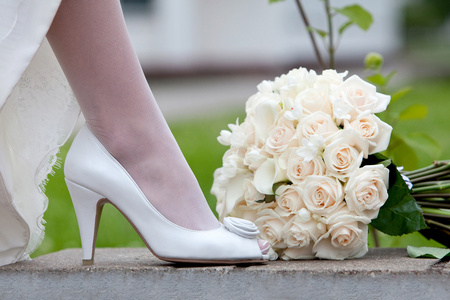 婚礼鞋和新娘花束。女性的脚，穿着白色的婚纱的鞋子和花束的新鲜的白玫瑰特写