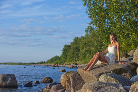 坐在湖岸上的岩石上的白色连衣裙的少女