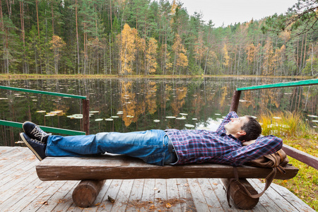 年轻男性徒步旅行者在秋天的树林湖边休息