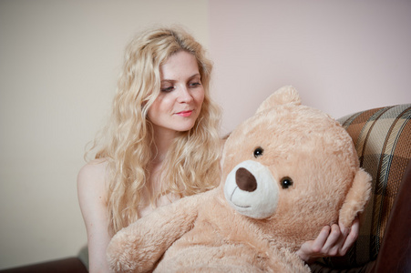 年轻的金发女人坐在沙发上放松与巨大的玩具熊。在沙发上用玩具放松的舒适的衣服的漂亮女孩。在室内舒适风景迷人金发女郎