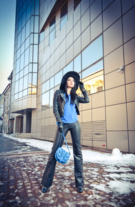 年轻漂亮的女人在冬季时尚拍摄。美丽时尚的年轻女孩，在黑色皮革与大的帽子和蓝色手袋前现代的建筑，城市风光的照片