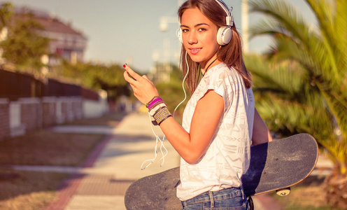 年轻女孩与滑板和头戴耳机收听音乐户外