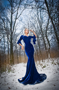 美丽的年轻女子的构成冬天的景色，看起来很高贵的优雅蓝色礼服。时尚的金发女人与森林在户外拍摄的背景下。迷人的金发女性性质的  