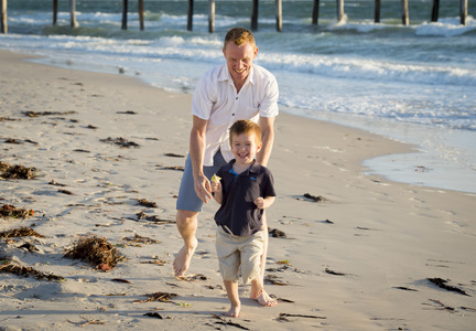 快乐爸爸玩在沙滩上与运行的小儿子兴奋与赤脚在沙子和水