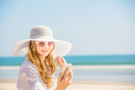 美丽的年轻女子在沙滩上坐在阳光明媚的日子，在她手中的手机