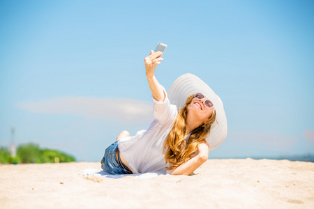 美丽年轻女子躺在沙滩上在阳光明媚的日子，以自拍照与电话