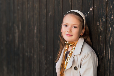 一个可爱的小女孩，穿风衣和金色围巾站在黑暗的木墙的室外肖像