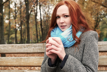 杯茶坐在城市公园的长凳上的红发女孩的秋天