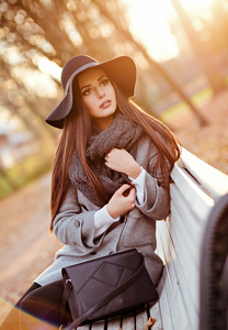 非常漂亮的年轻黑发女人灰色外套和黑色的帽子，坐在板凳上为背景，在公园里的秋景直头发光泽的肖像