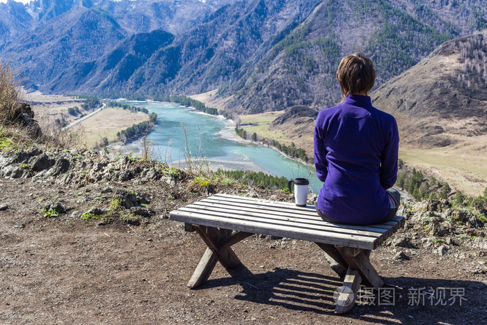 年轻女子坐在长椅上,看着一条河与一杯茶