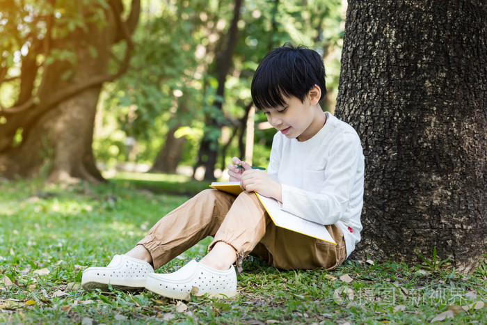 亚洲的小男孩,坐在树下和画在笔记本上