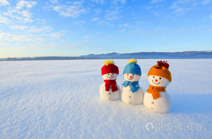 三个戴着五颜六色帽子的小雪人站在雪地上雪蓝色天空中有山野的风景