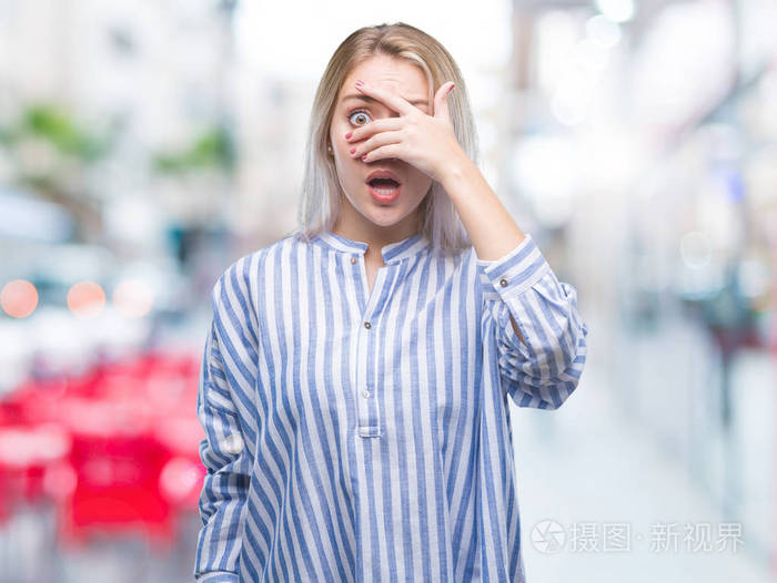 年轻的金发女子在孤立的背景下,震惊地偷看,遮住脸和眼睛,用手透过