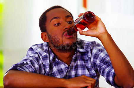 英俊的男人穿着白色蓝色衬衫坐在酒吧柜台旁，躺在桌子上，喝着棕色的啤酒瓶，喝醉了