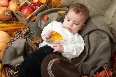 秋天的小男孩躺在黄色的落叶上，苹果，南瓜和纺织品上的装饰