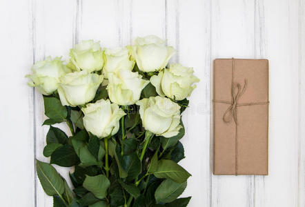 一束豪华的白色玫瑰和一个现在的盒子在生态纸上的白色木制背景。 俯视图，平躺