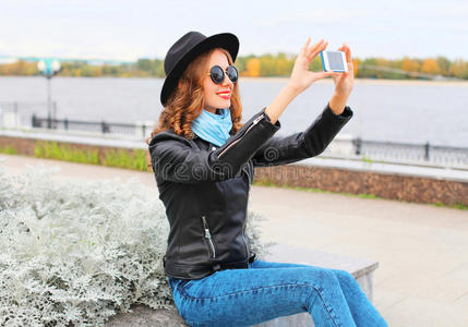 时尚酷的年轻微笑女孩在城市的智能手机上拍照自我肖像