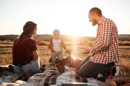 快乐的年轻家庭的妈妈和爸爸带着他们的小儿子享受着夏日周末的郊外野餐，在阳光明媚的夕阳下，享受假期的概念