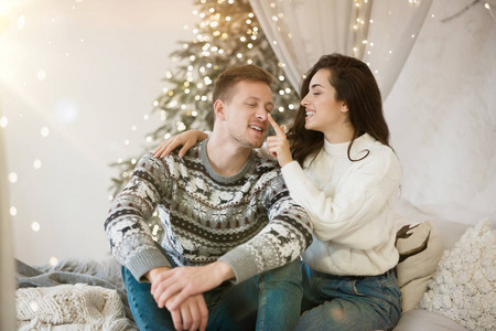 可爱的年轻夫妇漂亮的女人和英俊的男人穿着毛衣拥抱着鬼脸在沙发上开心地庆祝新年圣诞节的节日气氛